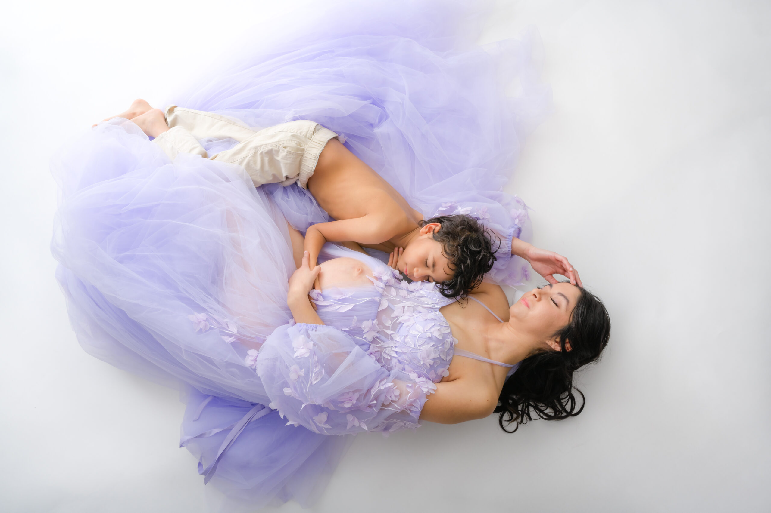 professional maternity photography of mom to be and son snuggling together laying on a white backdrop.