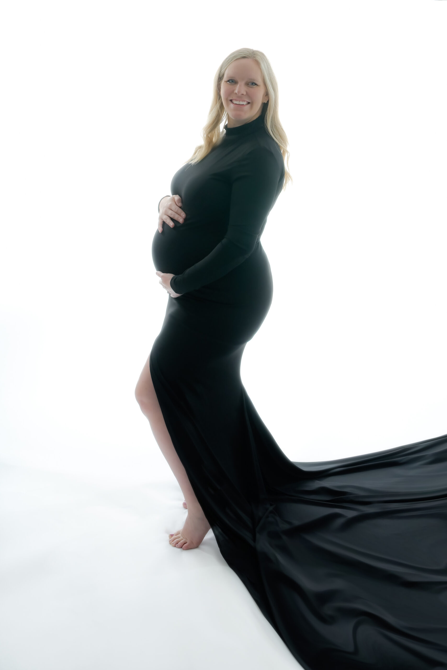 Backlit maternity photo session with mom to be wearing a long black gown with a train spread out.
