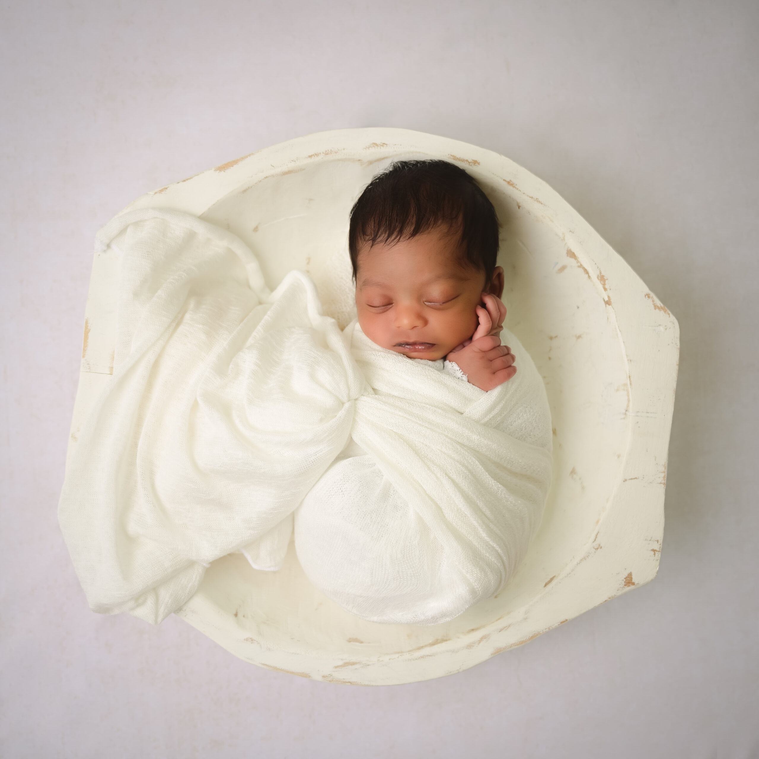 Newborn boy wrapped in cream colors posed sleeping in a cream bowl